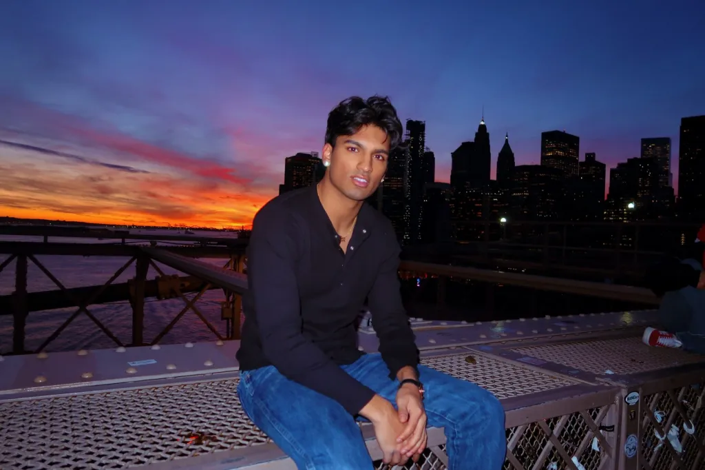 Jasik Dhillon sitting on a bridge with a sunset and city skyline in the background.