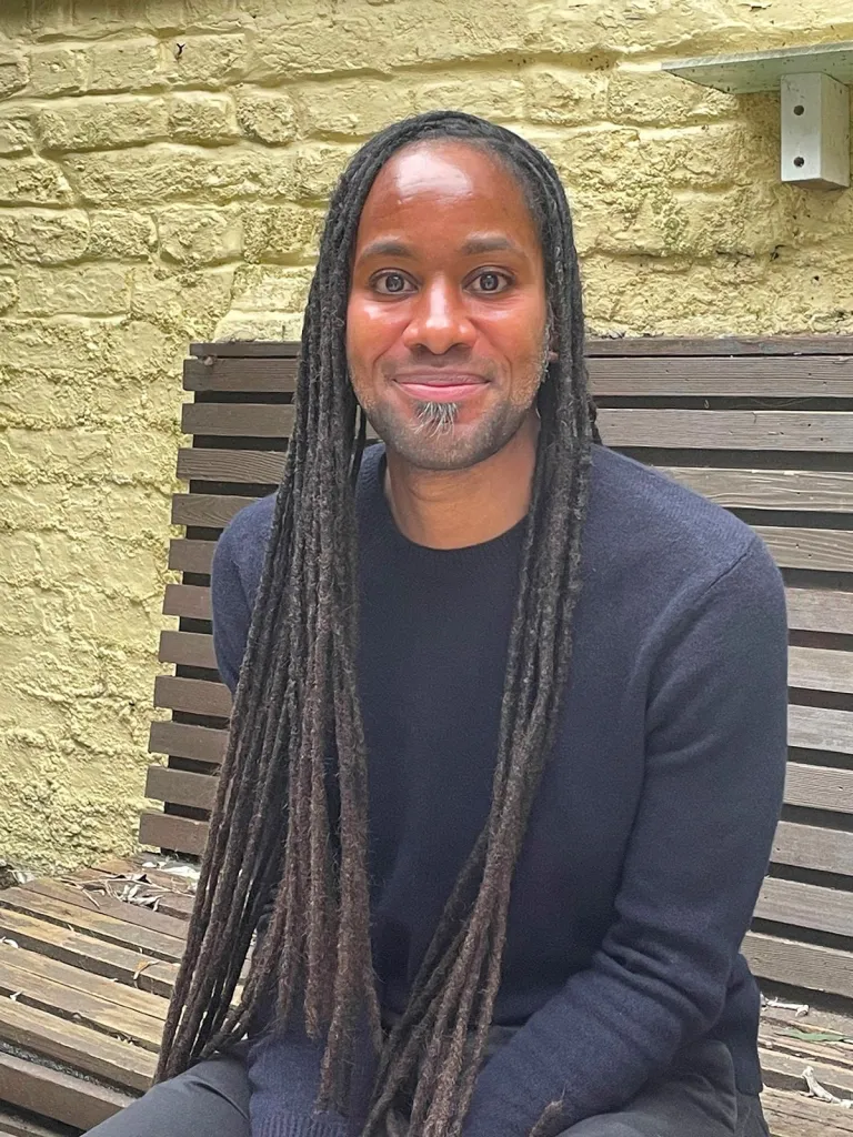 Jason Arday, a Black man with dreadlocks and a light beard, smiles while sitting on a wooden bench against a yellow brick wall.