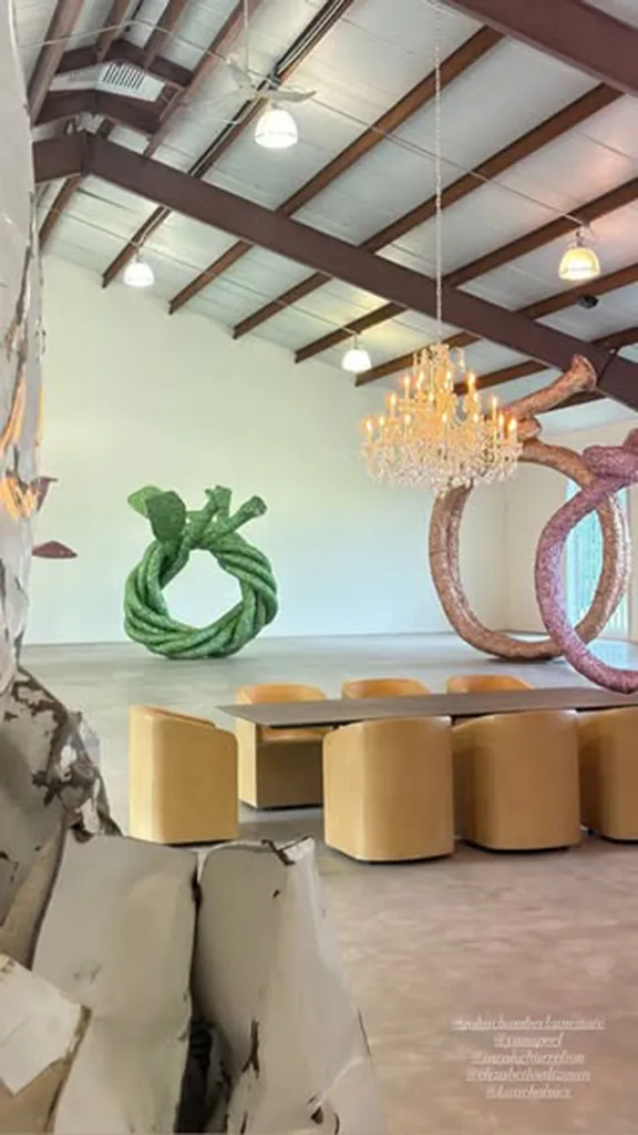 Room with a table and chairs, a green twisted rope sculpture, and a pink curved sculpture, under a chandelier and industrial ceiling.