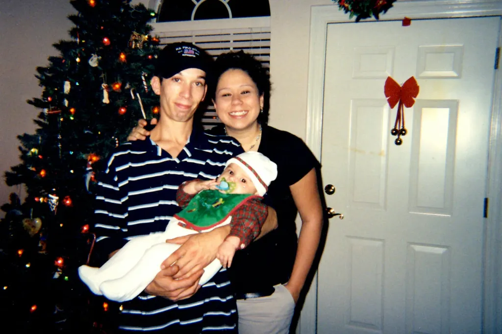 Jason Christopher Coulthart, wearing a striped shirt and a baseball cap, holding a baby, stands next to a woman near a Christmas tree.