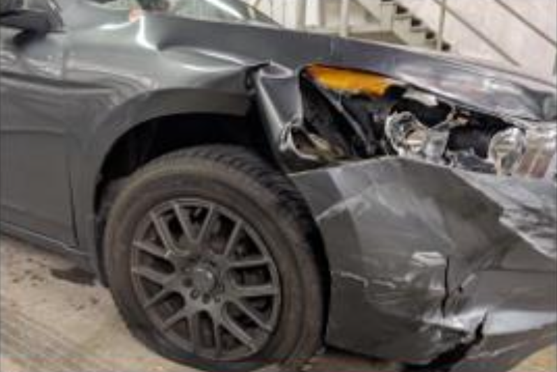 The front side of a dark gray car is heavily damaged, with the bumper and headlight smashed and the fender dented.