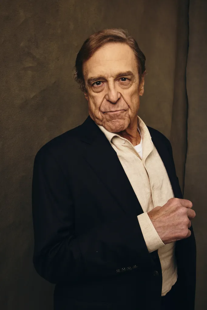 John Goodman in a dark suit jacket and cream-colored collared shirt, with a slight smile.