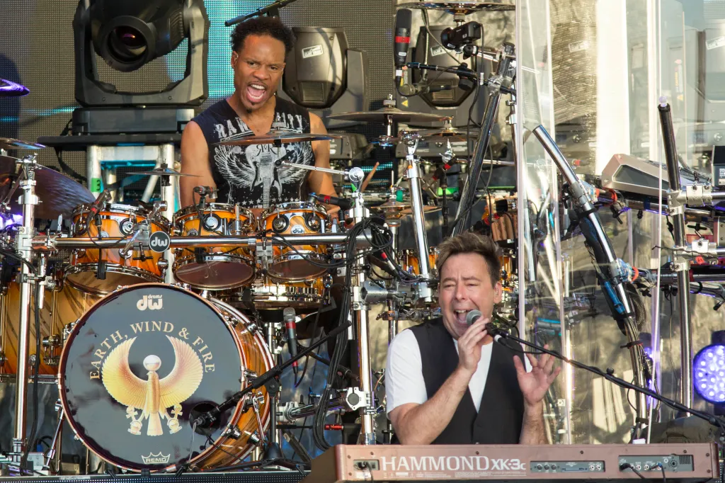 John Paris of Earth, Wind & Fire playing drums, and Lou Pardini of Chicago singing while playing keyboard at a concert.