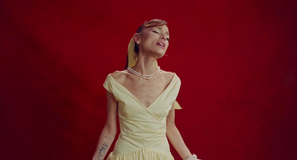 Ariana Grande in a yellow dress and pearls, singing with her head tilted back.
