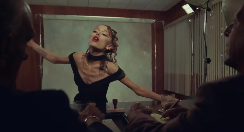 Ariana Grande performing "petal" in a black dress, leaning back on a table in front of two men.