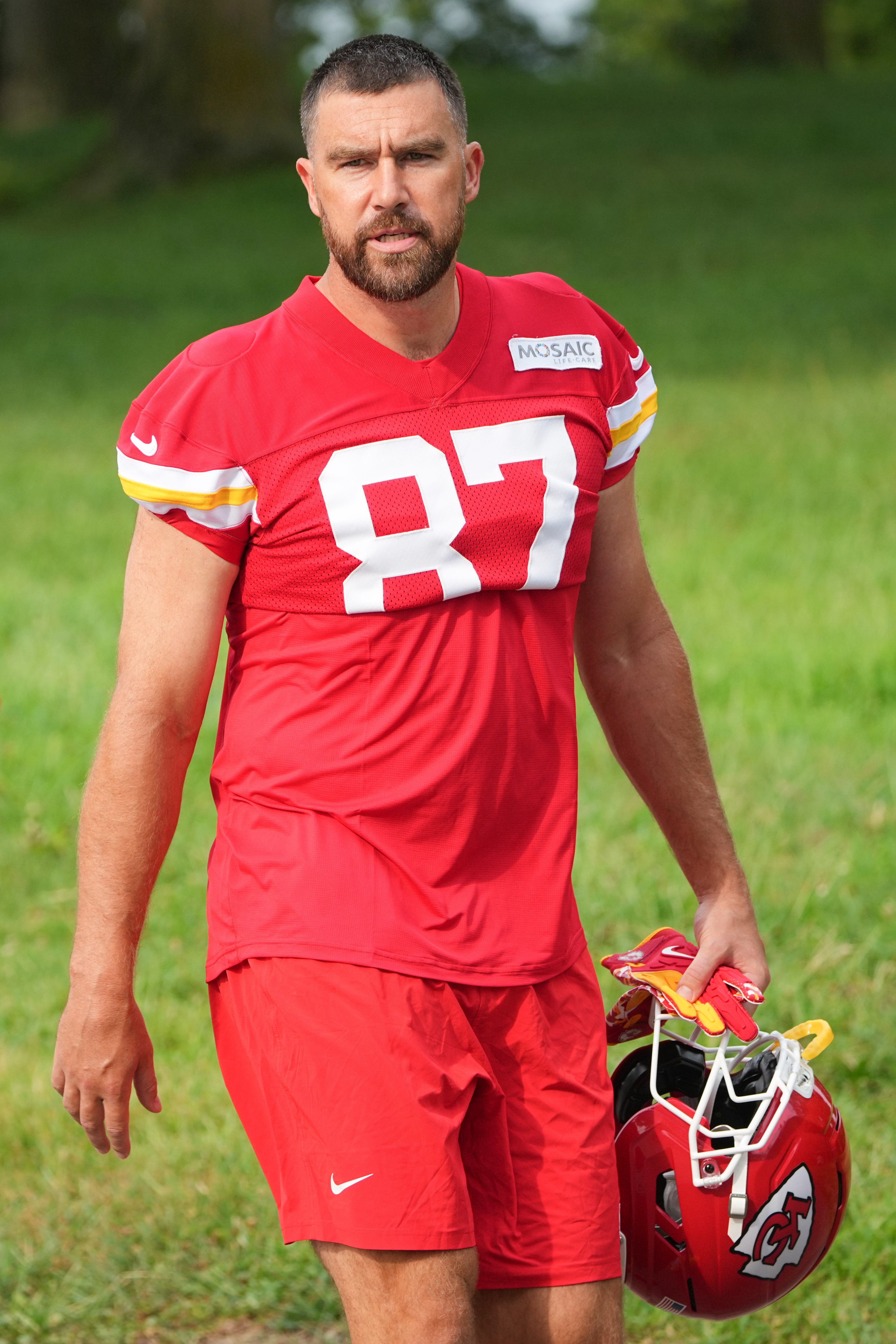 Kansas City Chiefs tight end Travis Kelce wearing a red practice jersey with number 87, holding a red helmet and gloves.