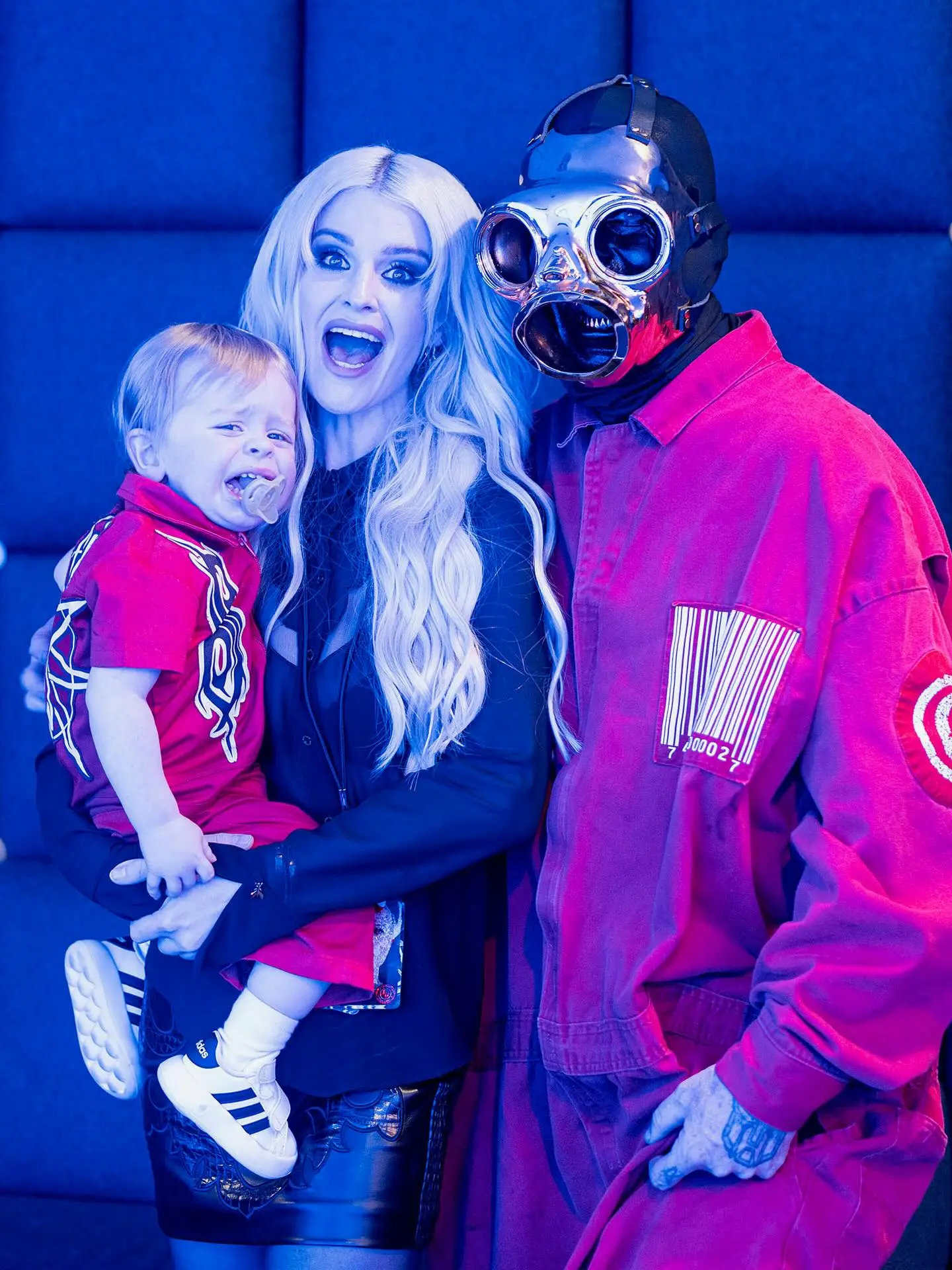 Kelly Osbourne holding her crying baby, next to Sid Wilson in a mask and red jumpsuit.