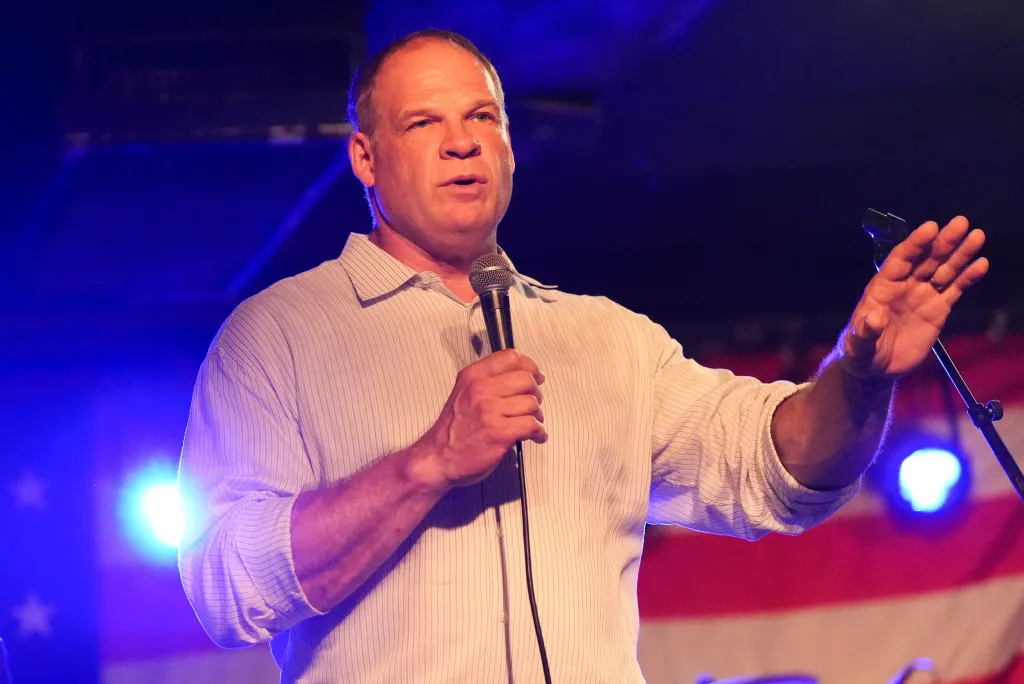Knox County Mayor Glenn Jacobs speaking at an event, holding a microphone, with a U.S. flag visible in the background.