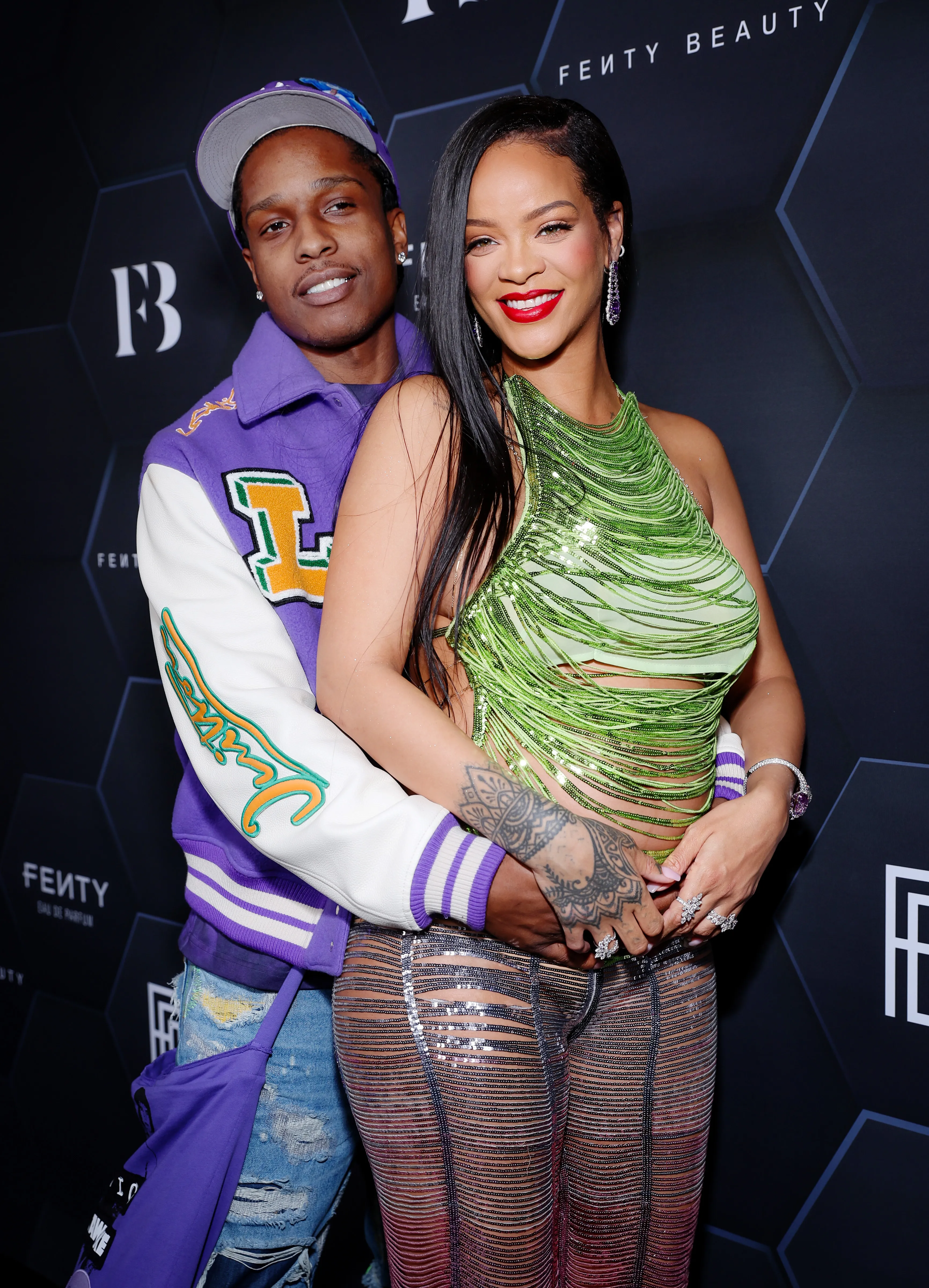 A$AP Rocky, wearing a purple and white letterman jacket, hugs Rihanna, who is in a shimmering green top and sheer, sequined pants.