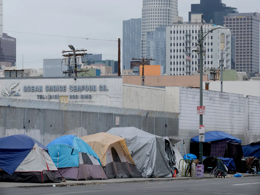 Homeless tents line a street in Skid Row, Los Angeles, with downtown buildings in the background.