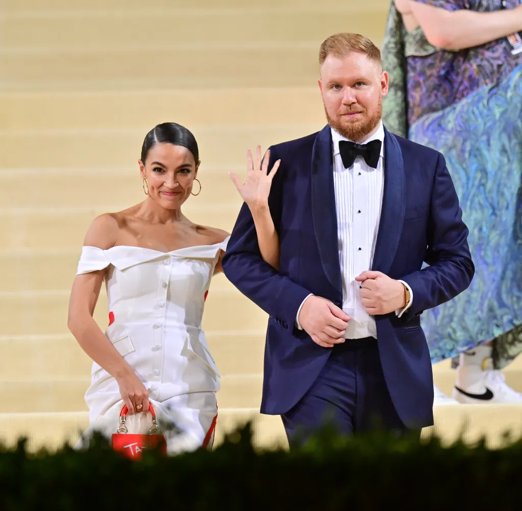 Alexandria Ocasio-Cortez in a white off-the-shoulder dress with a red handbag, holding onto Riley Roberts' arm, who is in a navy blue tuxedo.