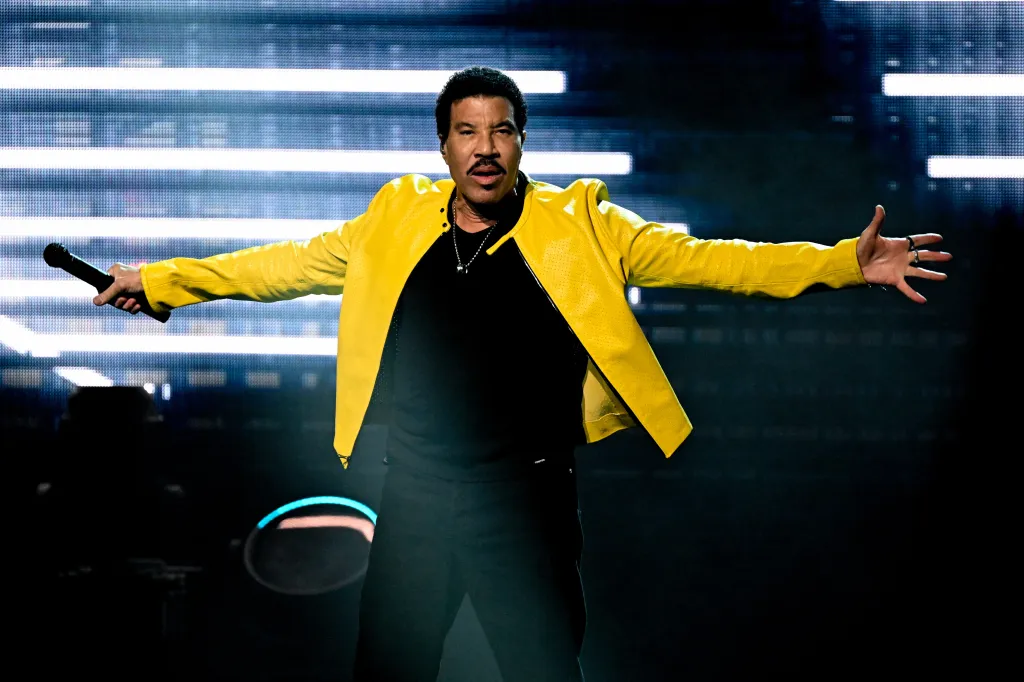 Lionel Richie performing on stage, wearing a yellow jacket and holding a microphone.