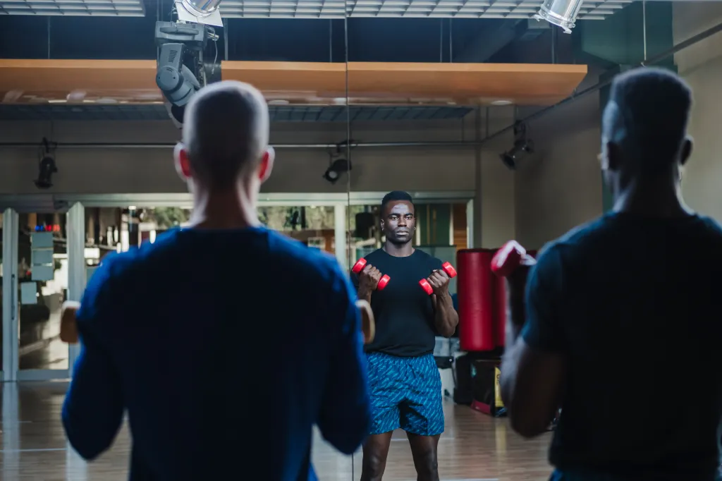 Two men exercising with dumbbells in a gym, seen through a mirror.