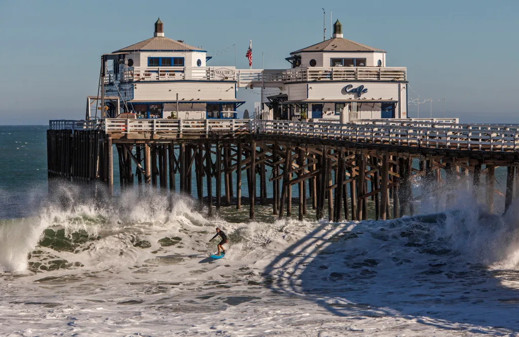 A surfer on a blue surfboard rides a large breaking wave underneath the Malibu Pier.