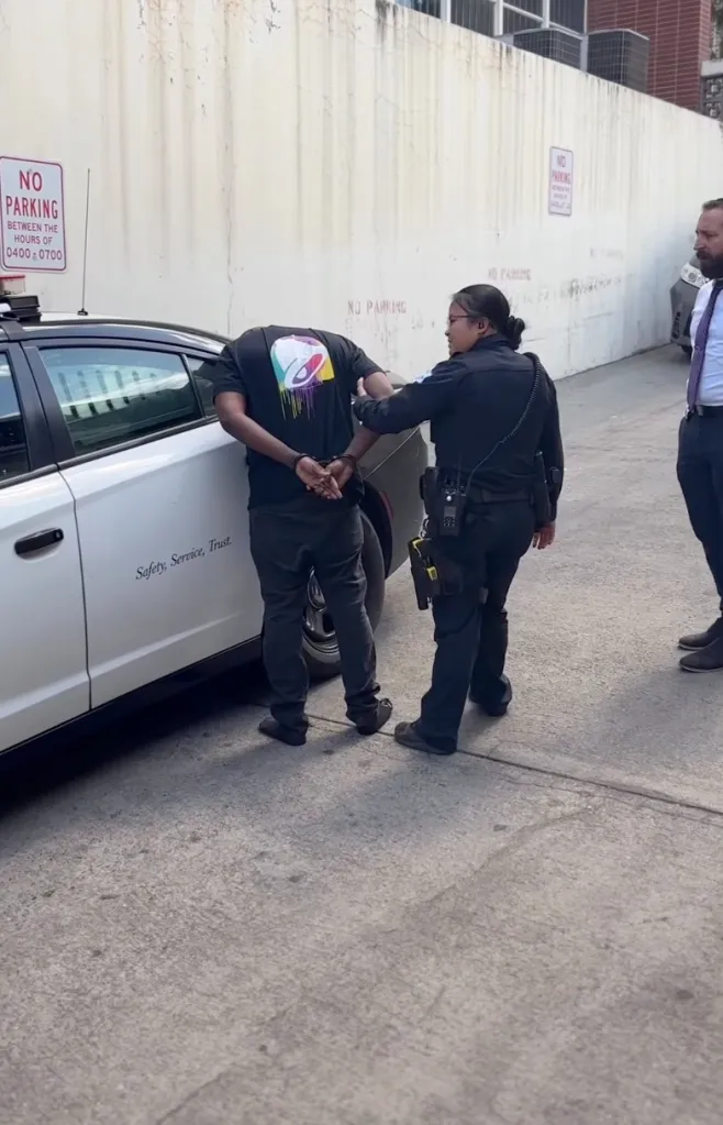 Man in handcuffs being led to a police car by an officer, with a third person standing nearby.