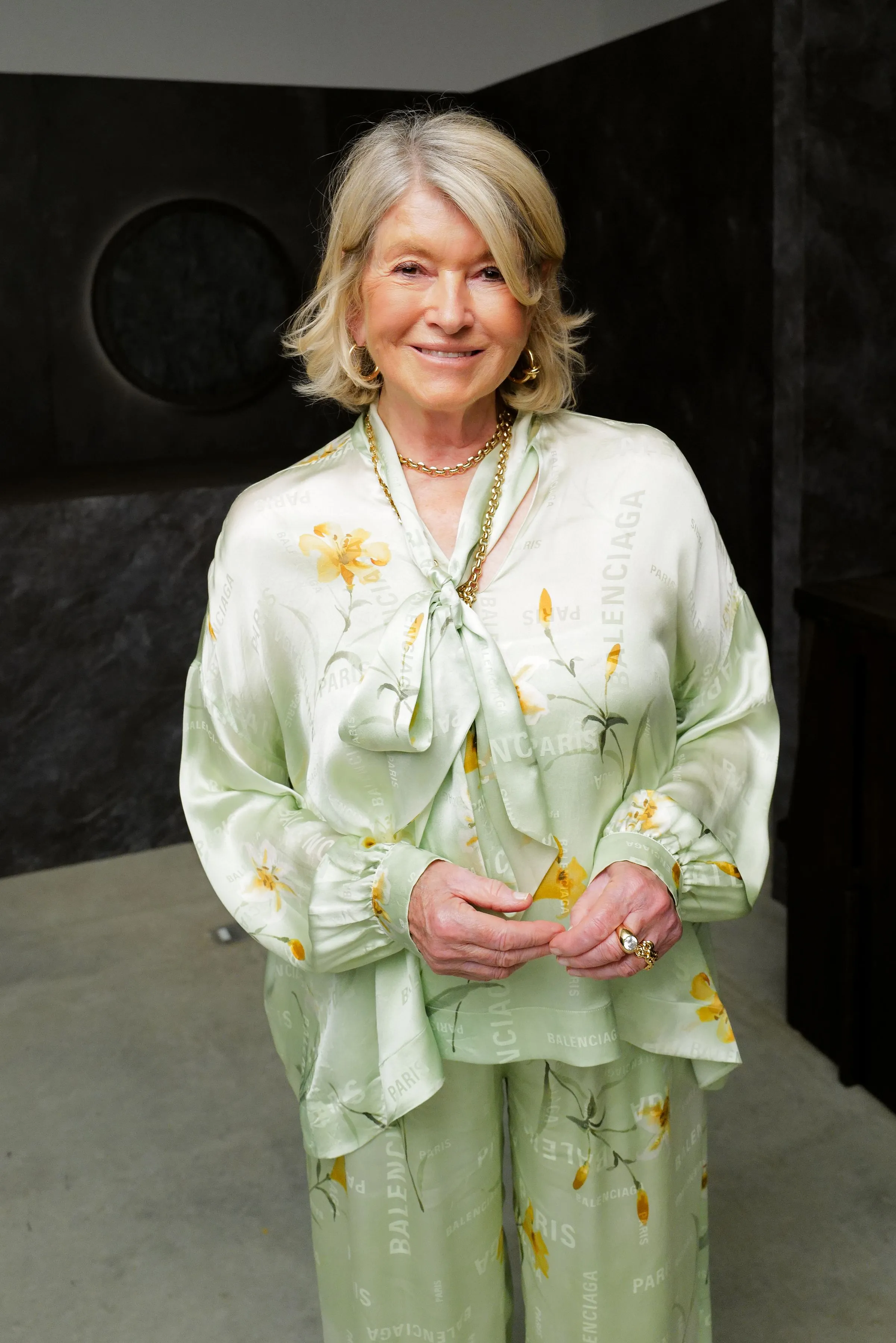 Martha Stewart smiling, wearing a light green satin blouse and matching trousers with a yellow floral and "Balenciaga Paris" pattern.