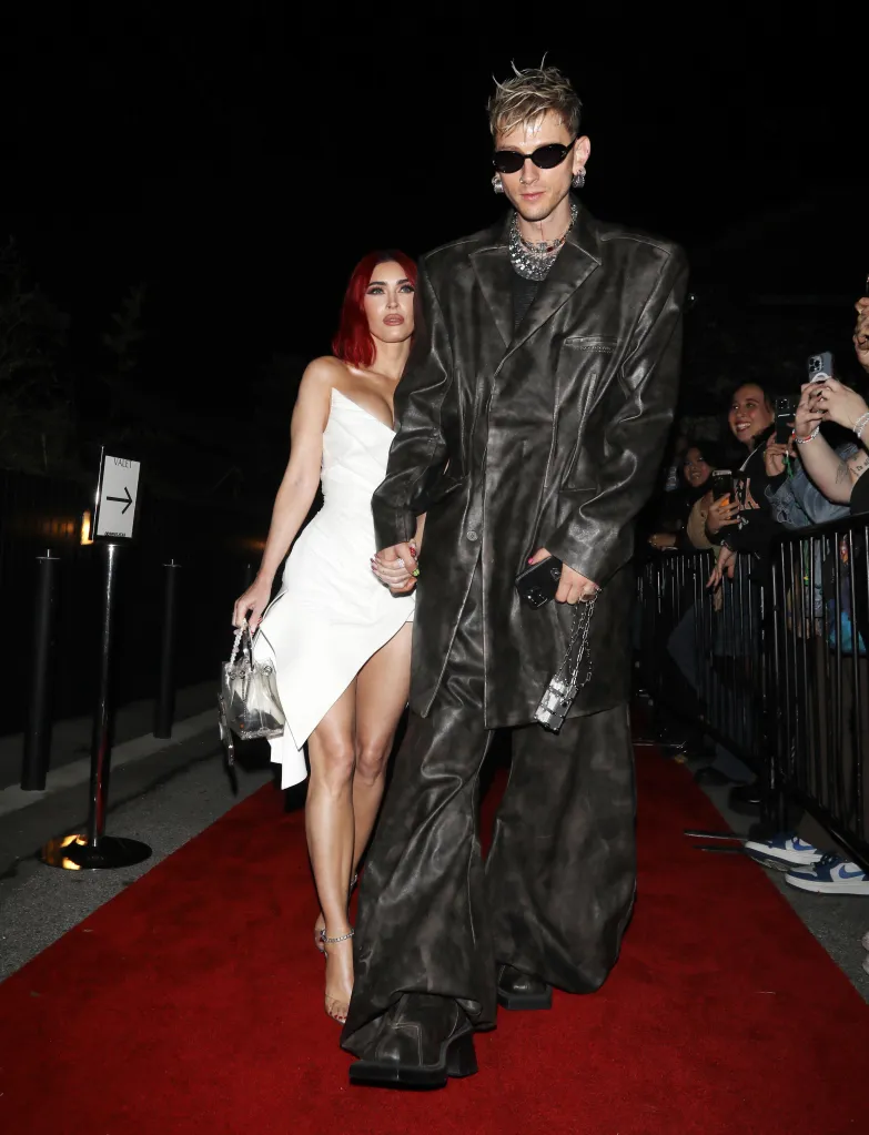 Megan Fox, in a white dress, and Machine Gun Kelly, in a large brown leather outfit, walk a red carpet.