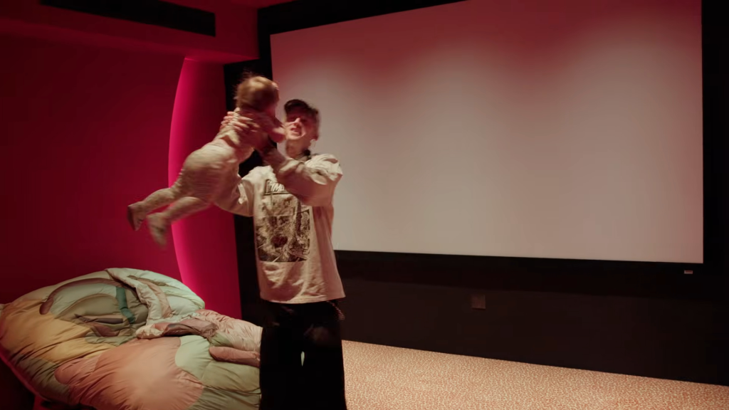 Machine Gun Kelly holding up his daughter, Saga, in a room with a large screen and red walls.
