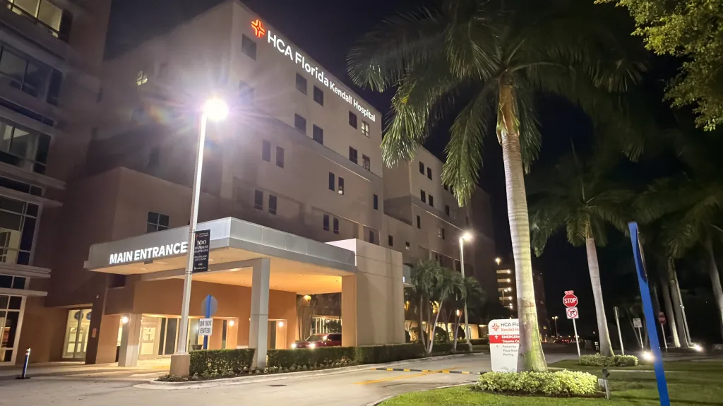 HCA Florida Kendall Hospital at night, illuminated by streetlights, with palm trees and a "MAIN ENTRANCE" sign.