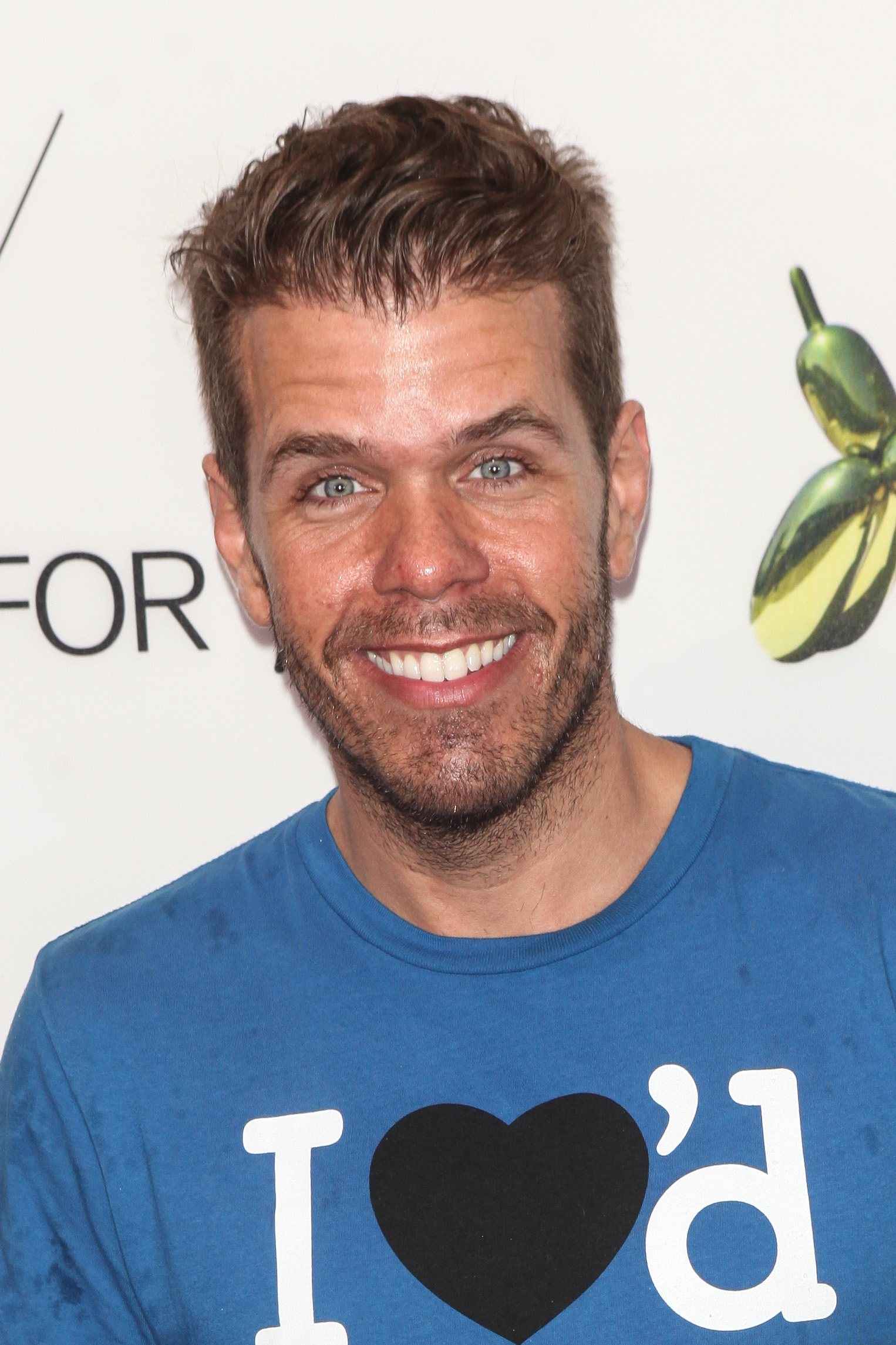 Perez Hilton smiling at the camera, wearing a blue t-shirt with a black heart and white text.