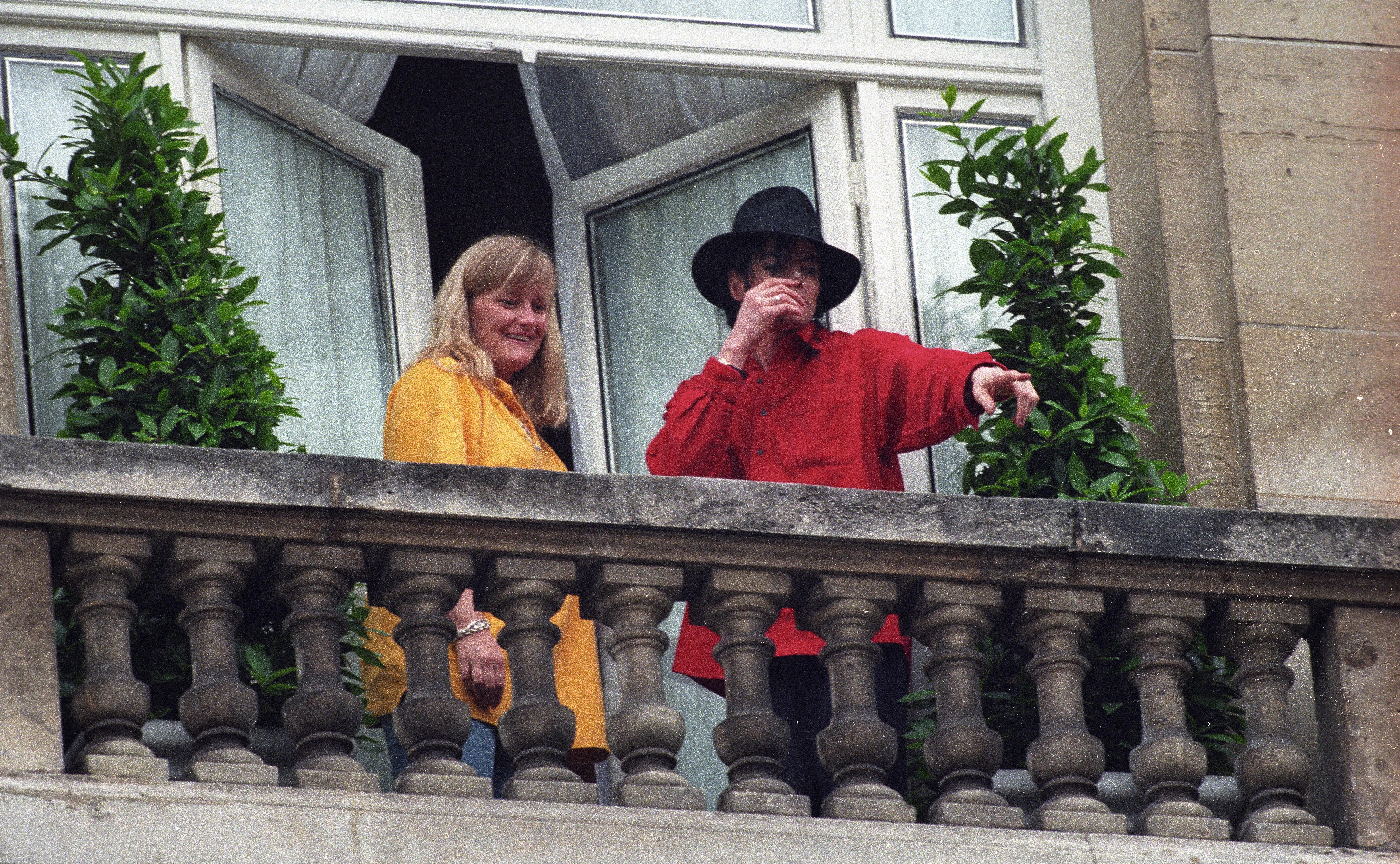 Michael Jackson, in a red shirt and black hat, and Debbie Rowe, in a yellow shirt, standing on a balcony.