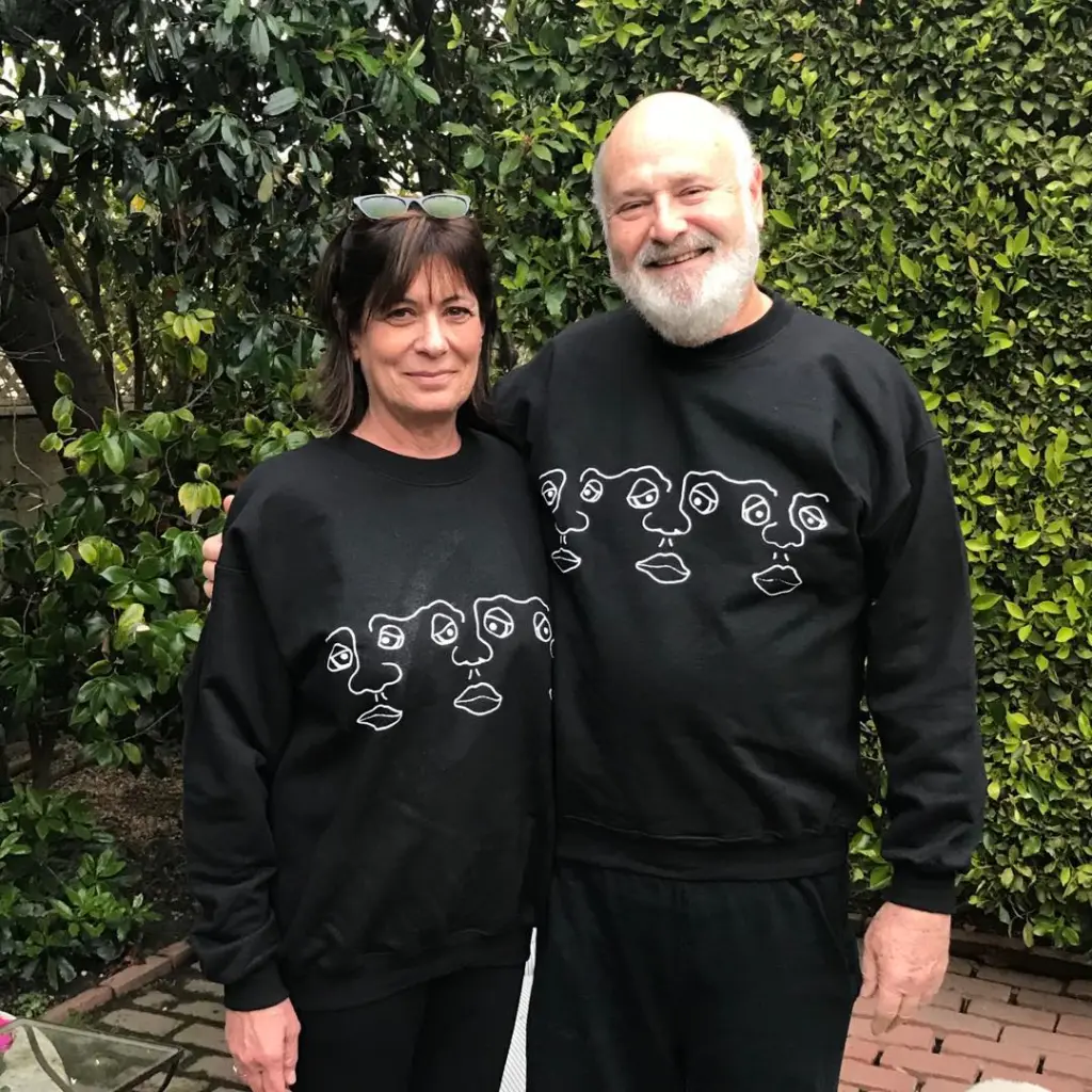 Michele and Rob Reiner smiling and wearing matching black sweatshirts with white line drawings of faces.