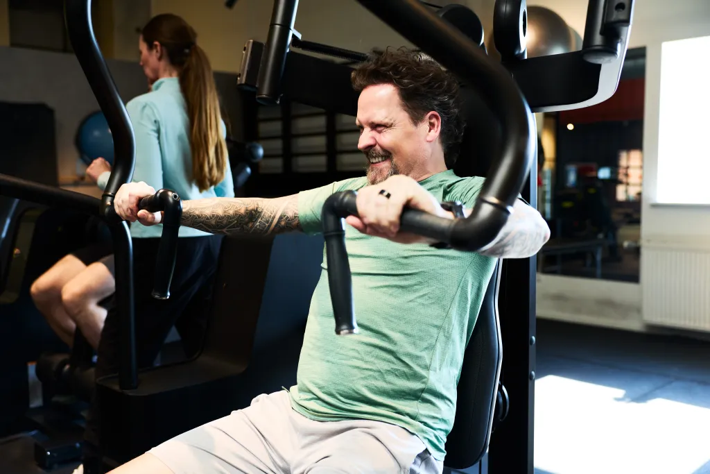 Man with tattoos on his arms, smiling with effort while using a chest press machine at the gym.