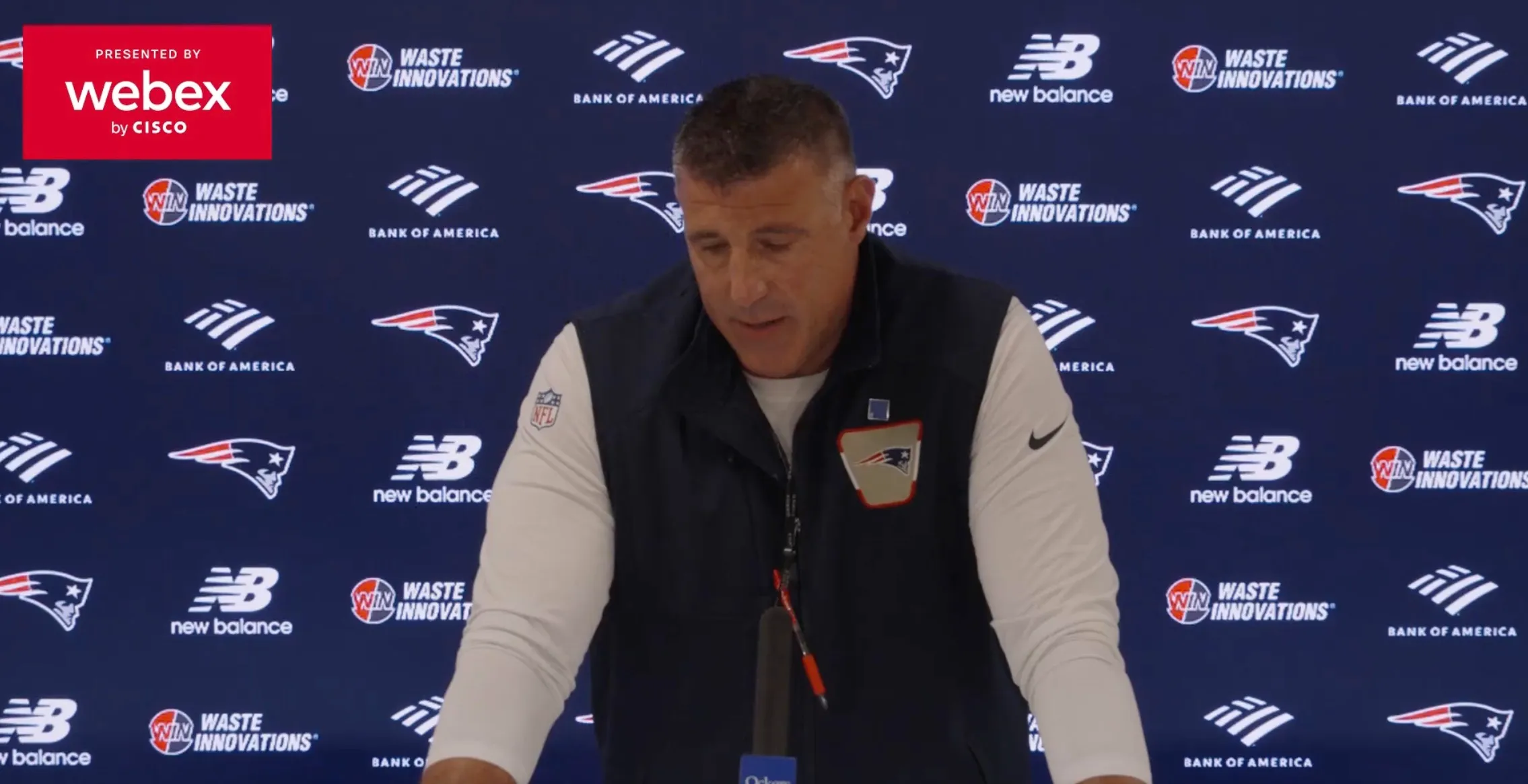 Mike Vrabel at a press conference with sponsor logos on the wall behind him.