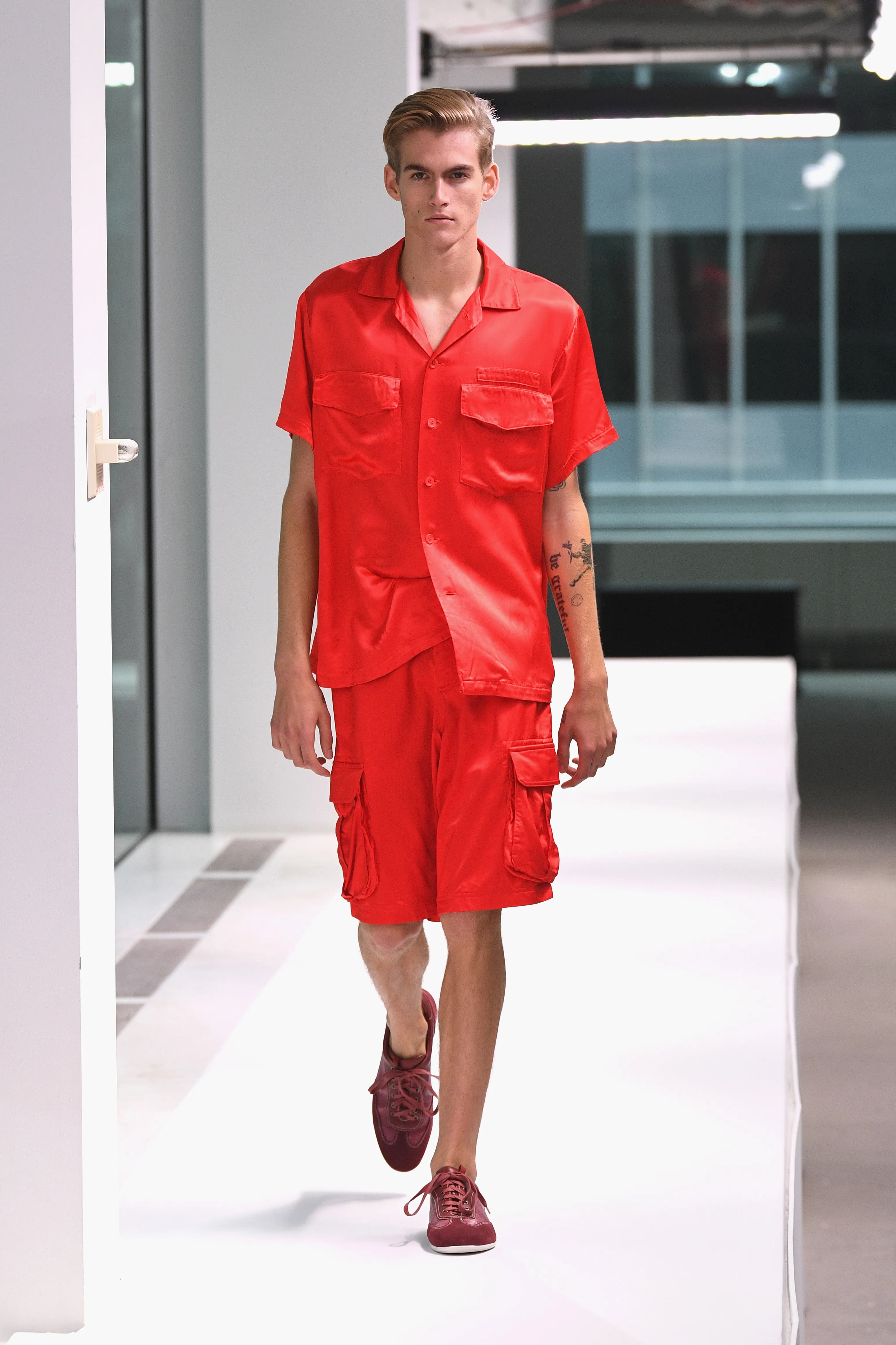 Presley Gerber walks the runway in a red satin short-sleeved shirt and matching red cargo shorts, with dark red shoes.