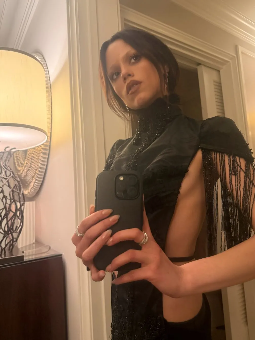 Jenna Ortega taking a mirror selfie in a black dress with beaded fringe.
