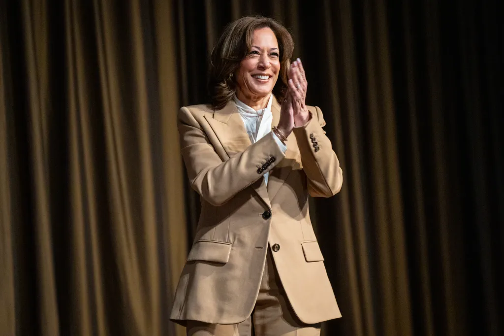 Kamala Harris, wearing a tan pantsuit and white blouse, smiles and claps her hands.