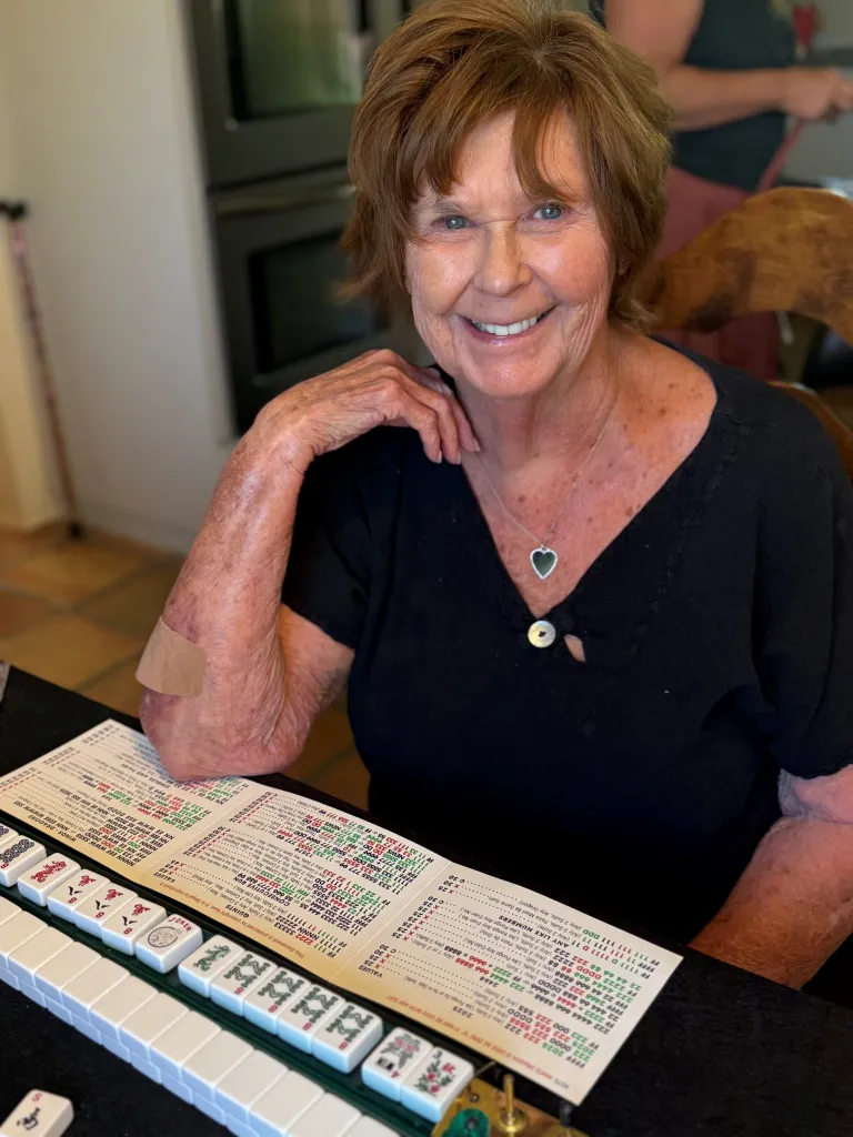 Nancy Guthrie smiling while seated at a table with Mahjong tiles and a game card.