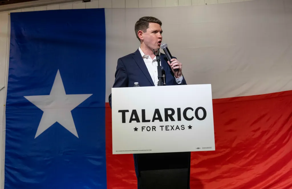 James Talarico speaking at a campaign rally in Lockhart, Texas, with the Texas flag in the background.