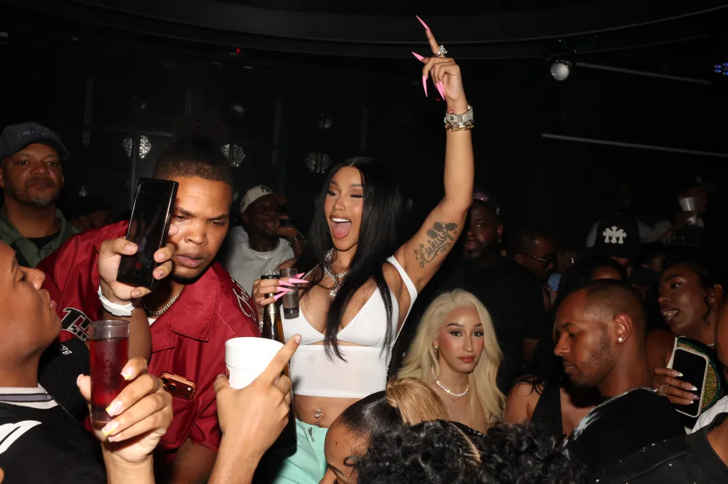 Cardi B in a white crop top and diamond jewelry, with pink stiletto nails, partying with a crowd.