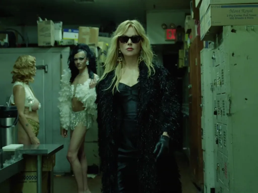 Nicole Kidman in sunglasses and a black feathery coat walks past two drag queens.