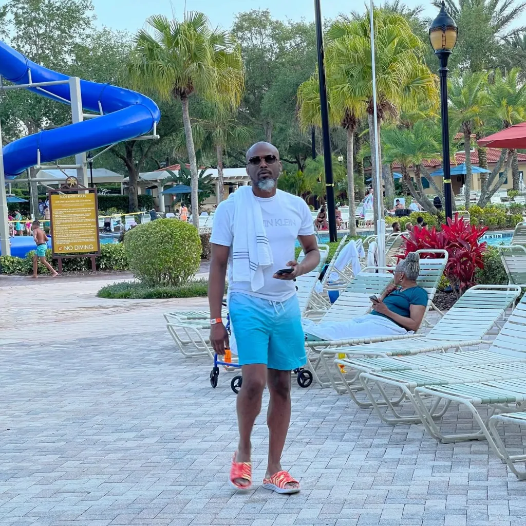 A man in a white t-shirt and blue shorts stands by a pool area with lounge chairs and palm trees.