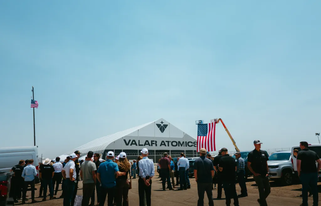 People gathered in front of a white tent with "VALAR ATOMICS" printed on it, with a large American flag hanging from a lift in the background.