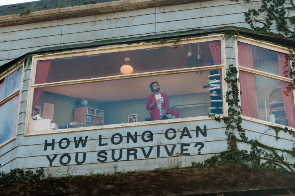 A man in a red robe sits in a fully furnished living room built into a billboard that reads, "HOW LONG CAN YOU SURVIVE?"