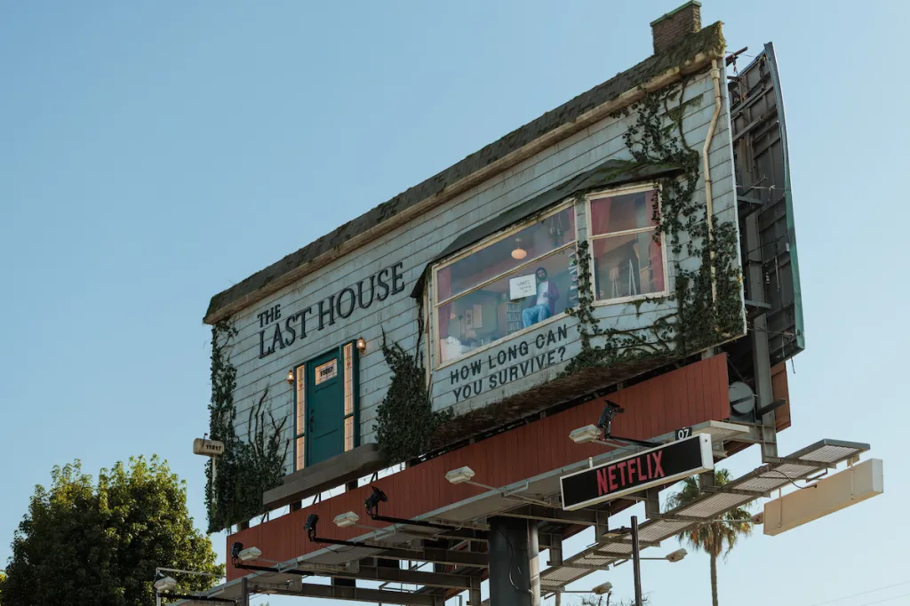 A man living in a furnished room built into a billboard advertising "The Last House" and Netflix.