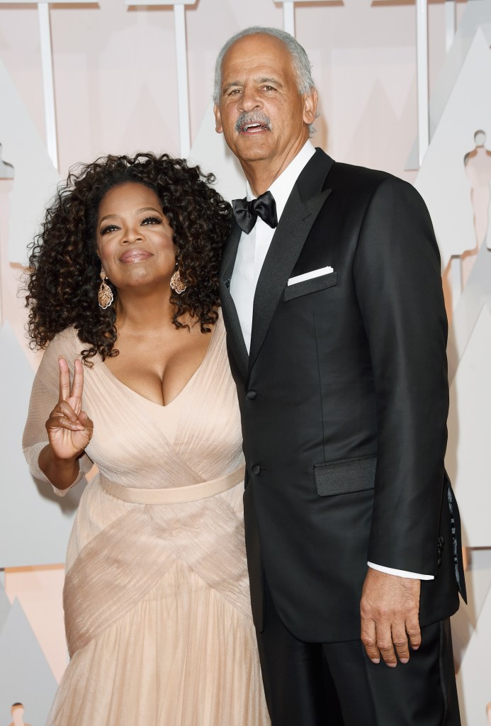 Oprah Winfrey in a pale pink gown, making a peace sign, and Stedman Graham in a black tuxedo stand together.