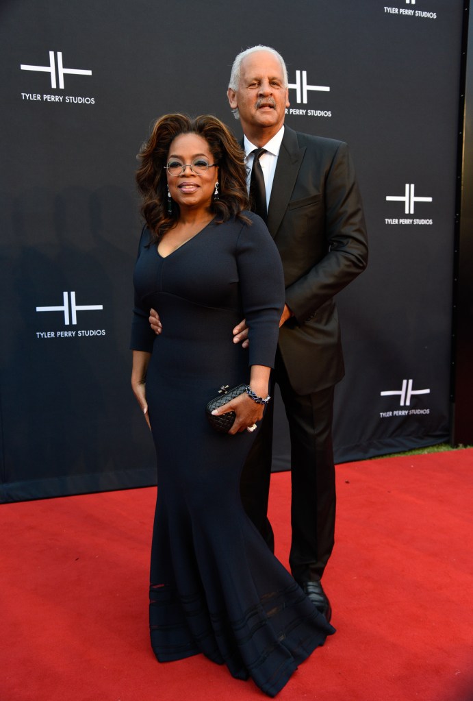 Oprah Winfrey in a black dress with Stedman Graham in a black suit, posing arm-in-arm on a red carpet in front of a black backdrop with the Tyler Perry Studios logo.