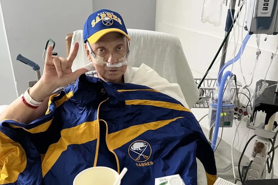 A patient in a hospital bed wearing a Buffalo Sabres hat and holding a Sabres jacket, making a hand gesture.