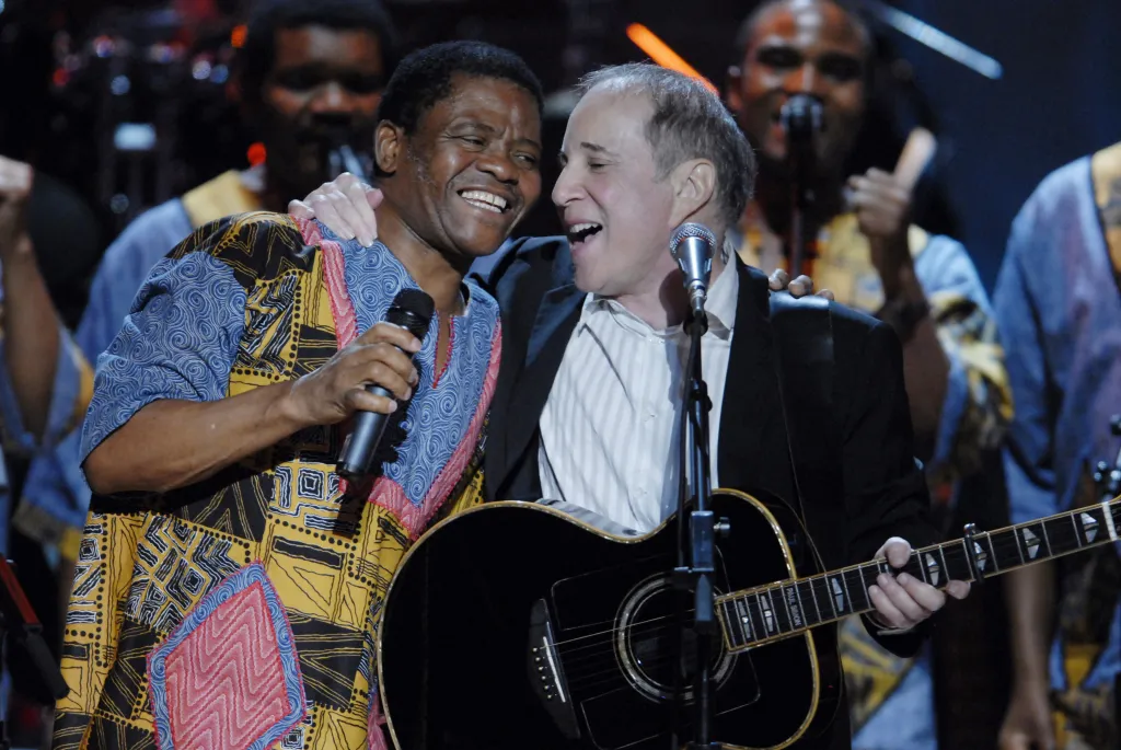 Paul Simon and a member of Ladysmith Black Mambazo laughing together on stage, Simon holding an acoustic guitar.