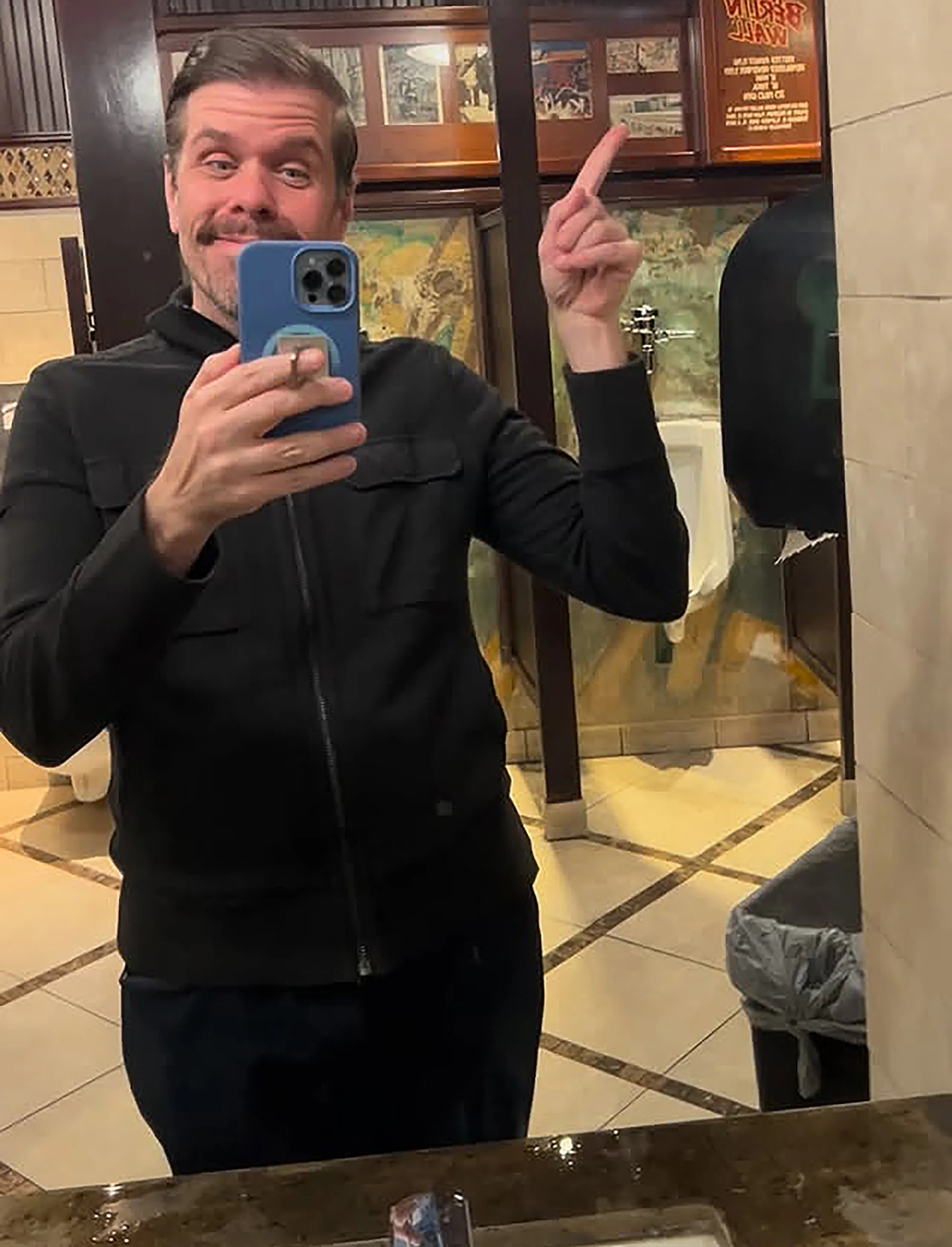 Perez Hilton takes a mirror selfie in a restroom, pointing upwards.