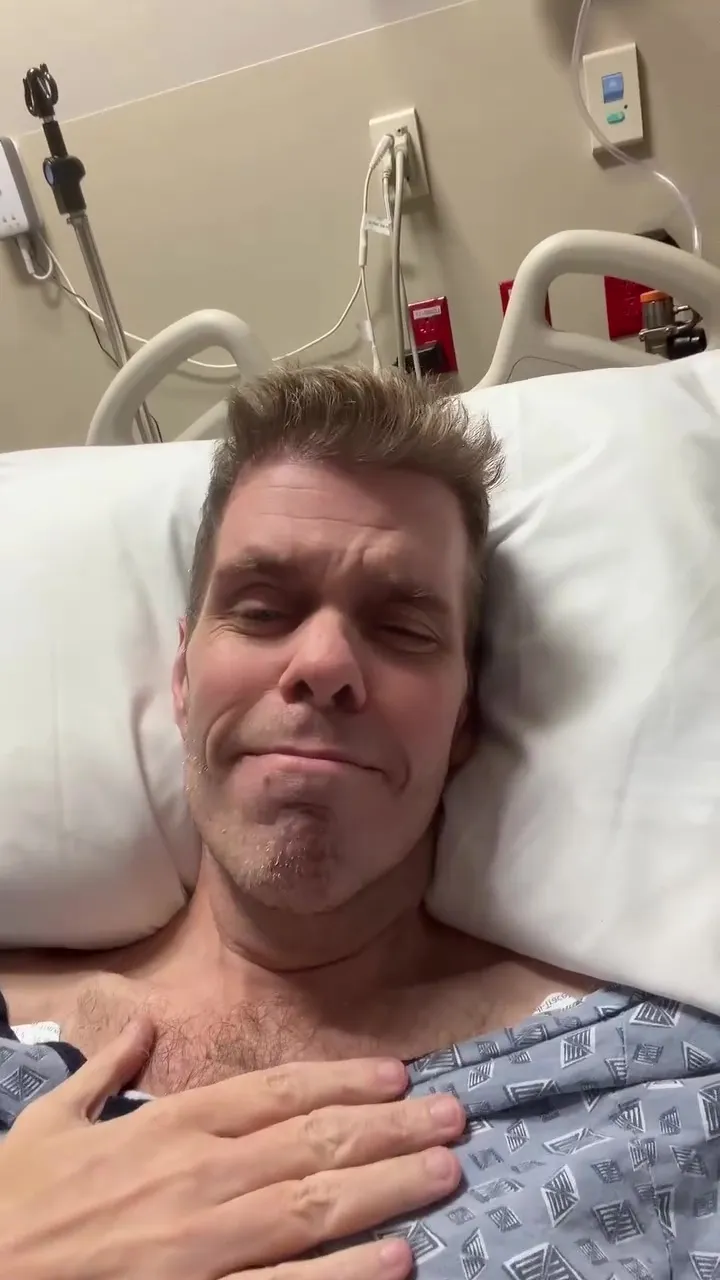 Perez Hilton lying in a hospital bed wearing a patterned gown.