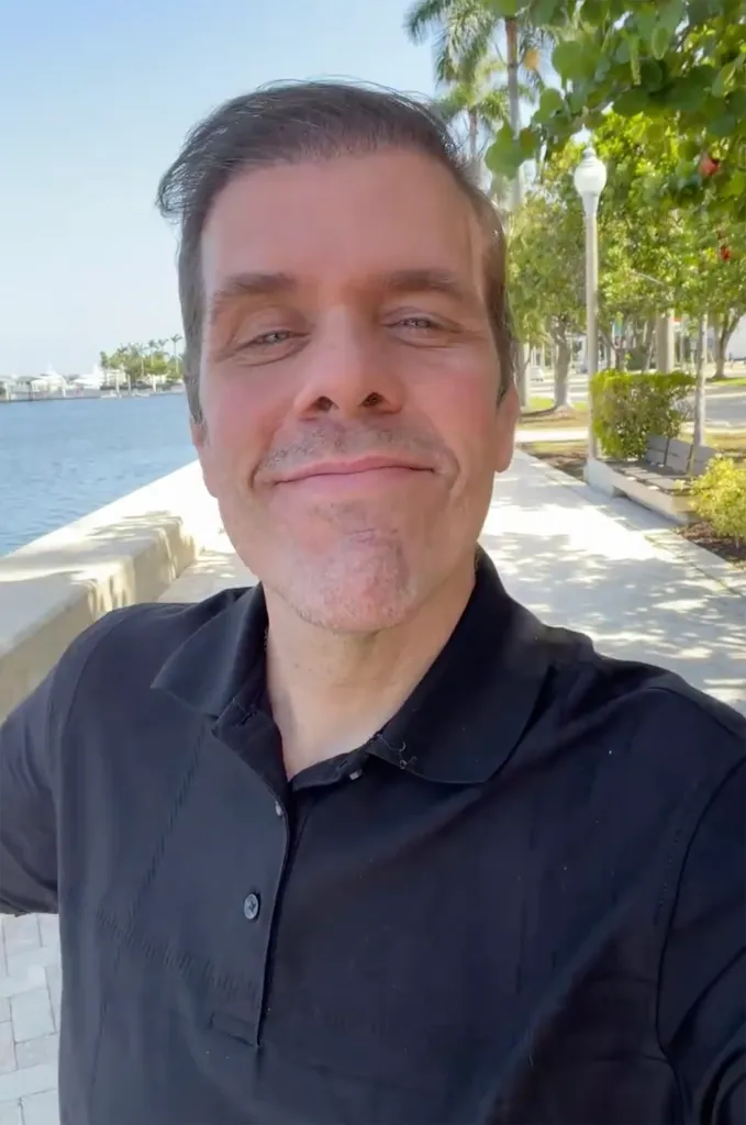 Perez Hilton smiling while standing outdoors next to a body of water.