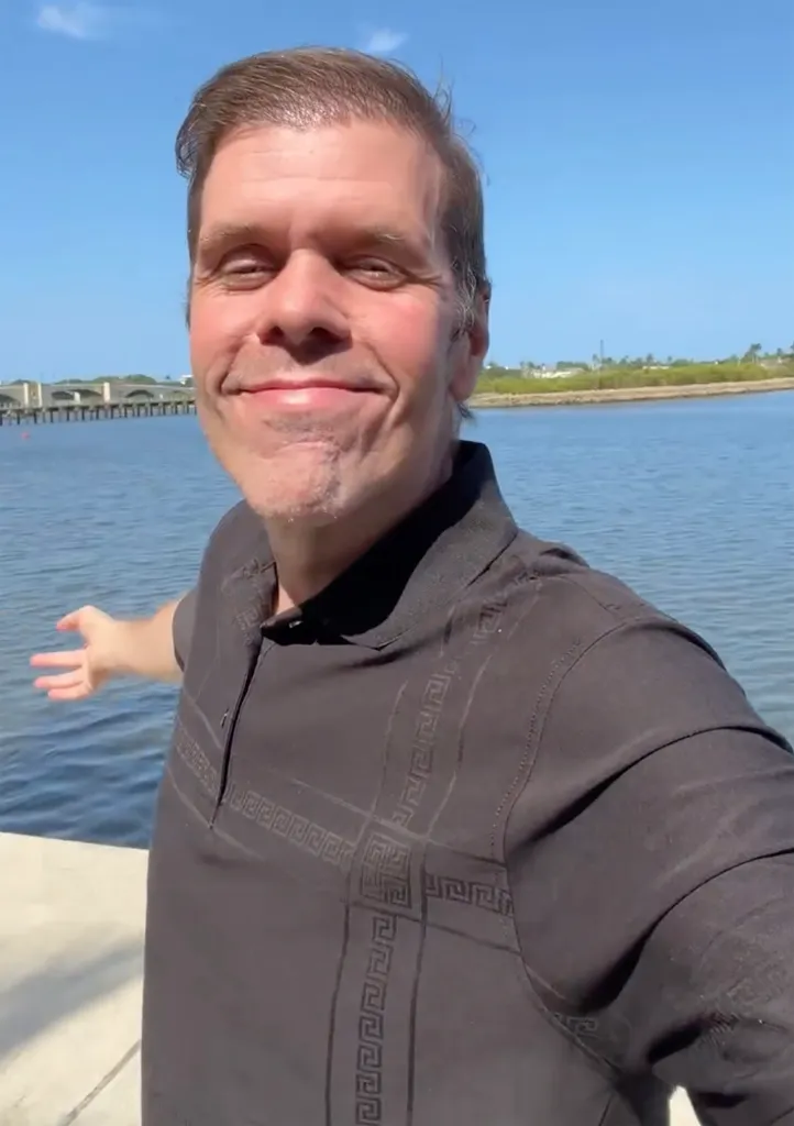 Perez Hilton smiles at the camera with a body of water and bridge in the background.