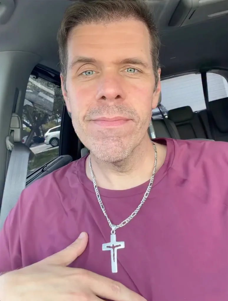 Perez Hilton wearing a plum shirt and a silver crucifix necklace, sitting in a car.