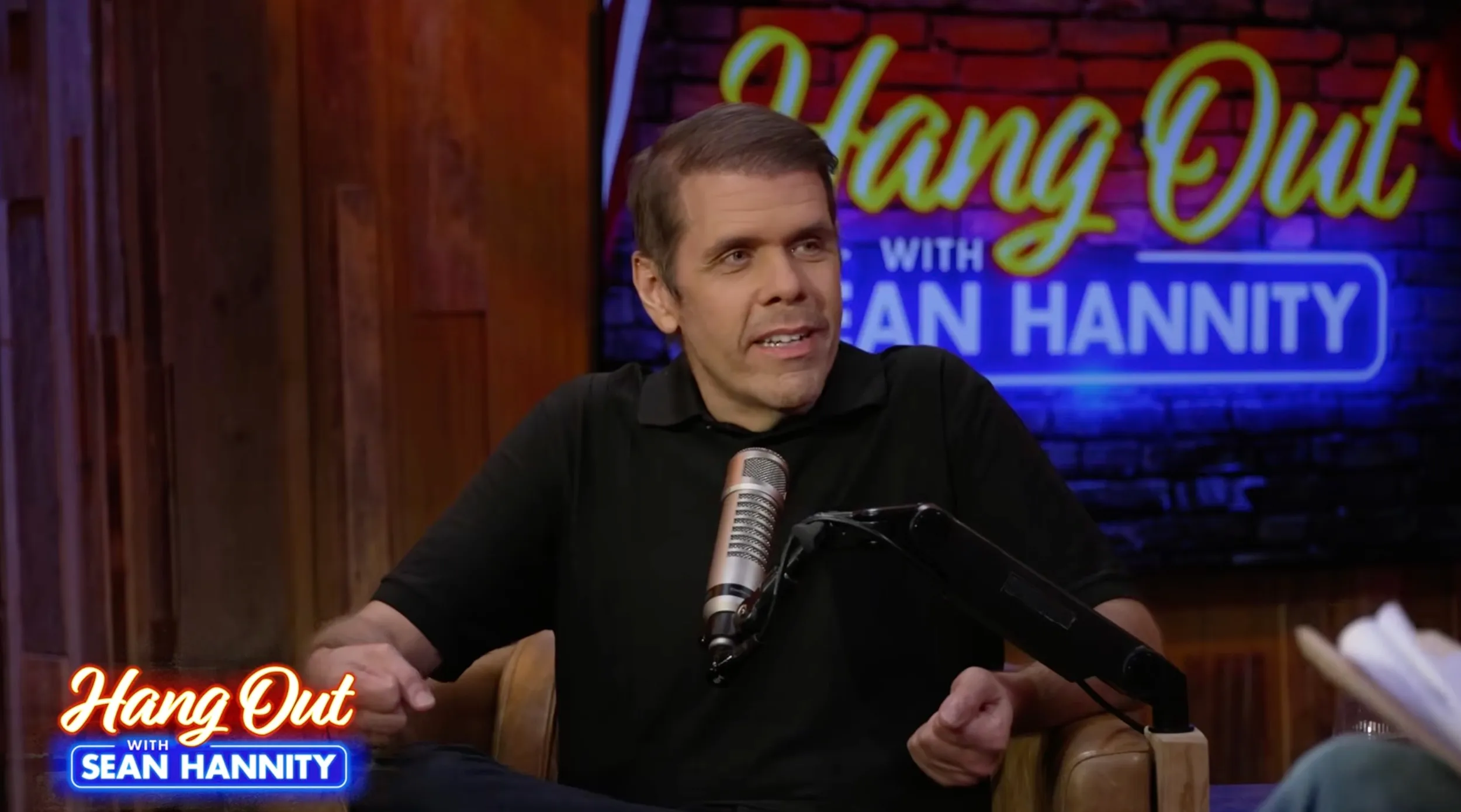 Perez Hilton wearing a black polo shirt while speaking into a microphone.