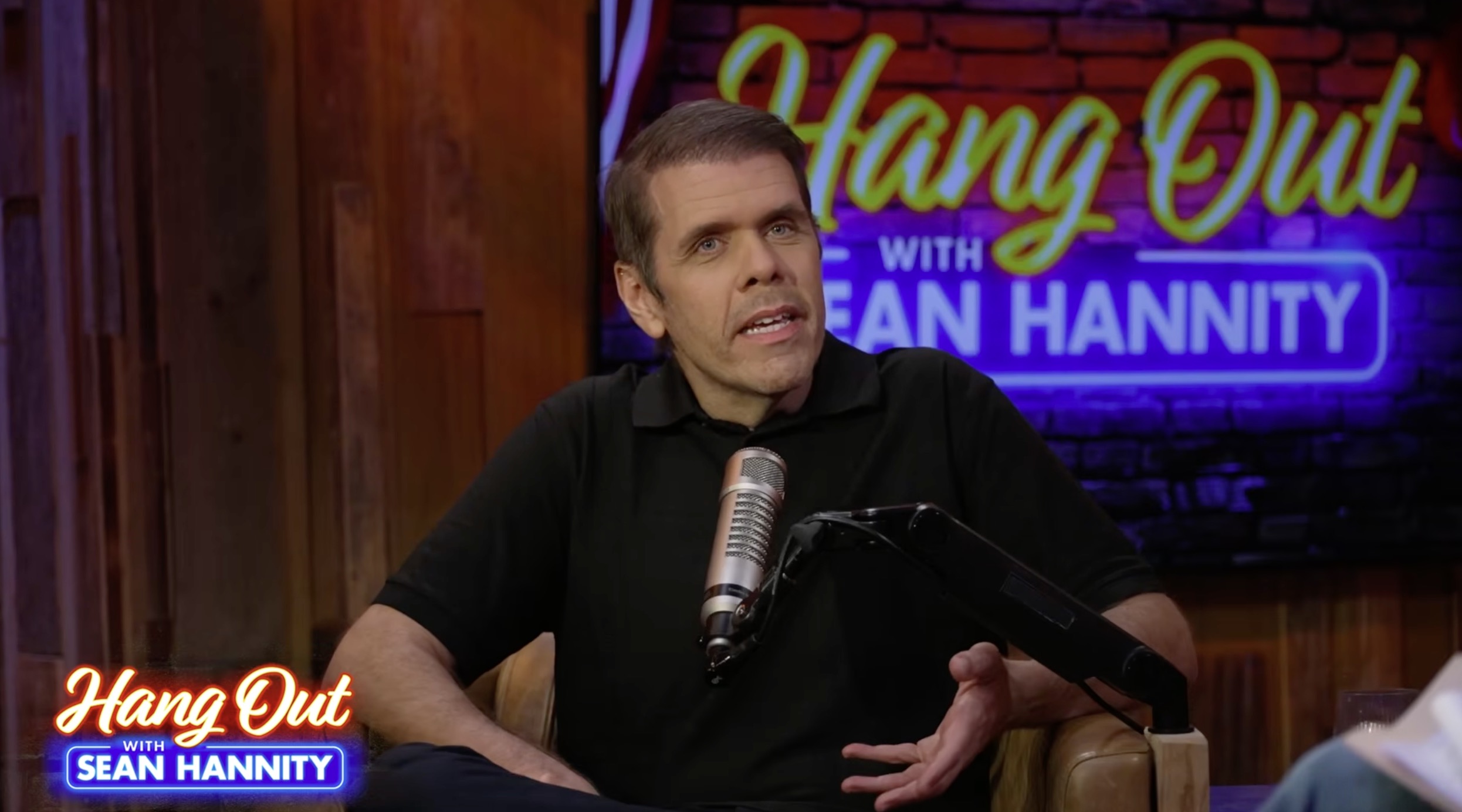 Perez Hilton in a black polo shirt, sitting next to a microphone with a neon sign that says "Hang Out with Sean Hannity" in the background.